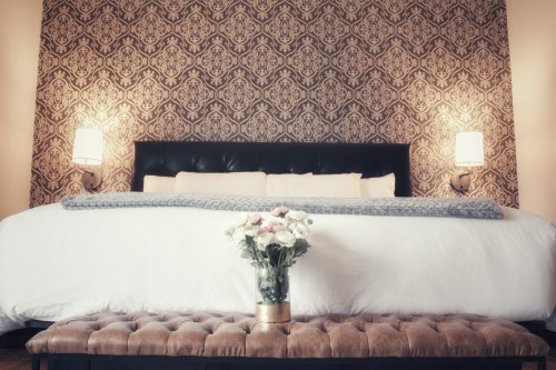 Cozy bedroom featuring a large bed with decorative pillows, a patterned wall, and a vase of flowers on a bench.