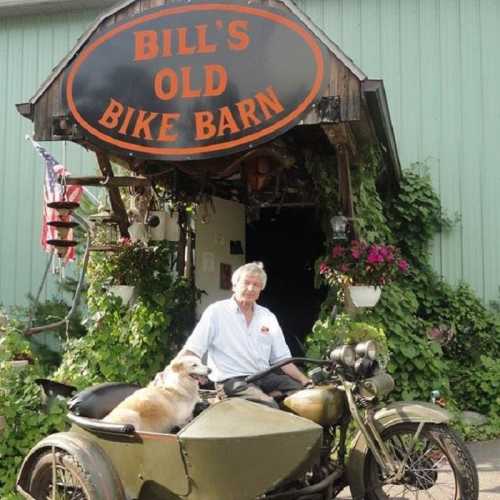 A man sits on a vintage motorcycle with a sidecar outside a rustic bike barn, surrounded by greenery and flowers.