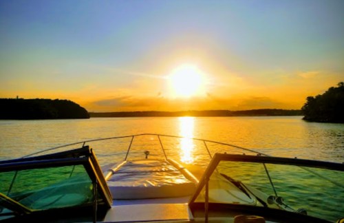A boat's bow facing a vibrant sunset over calm waters, surrounded by trees and hills.