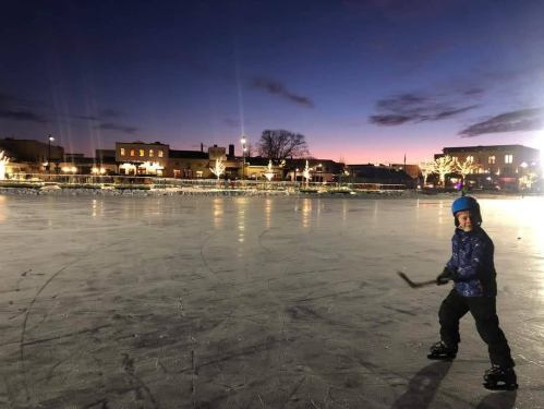 A child skating on an outdoor rink at dusk, with city buildings and lights in the background.