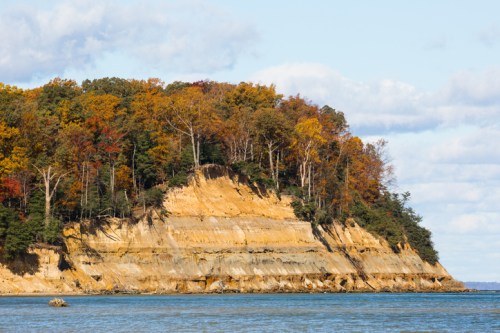A scenic view of a cliff with autumn foliage above and calm water below, under a partly cloudy sky.