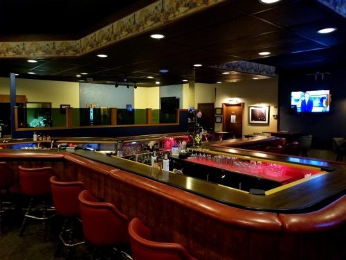 A dimly lit bar with a curved counter, red accents, and a TV in the background, featuring empty stools and shelves of drinks.