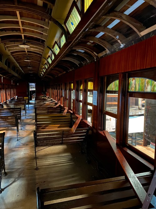 Interior of an old train car with wooden benches and large windows, showcasing vintage architecture and natural light.