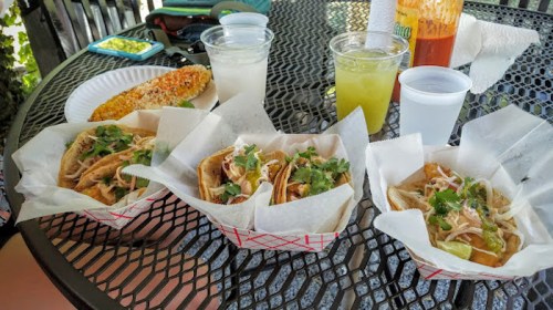 Three tacos topped with cilantro on a table, alongside drinks and a corn dish in the background.