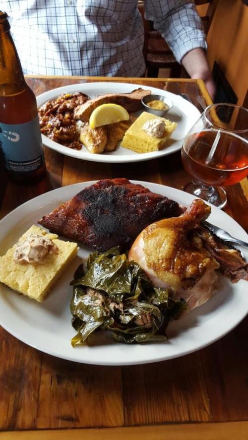 A plate of barbecue ribs, chicken, collard greens, cornbread, and a drink, with another plate in the background.