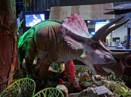 A colorful dinosaur model, resembling a triceratops, displayed in a themed environment with greenery and a warning sign.