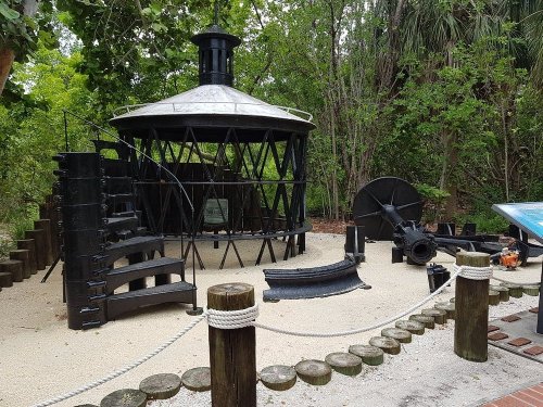 A historic lighthouse structure surrounded by trees, featuring a circular base and various metal components.