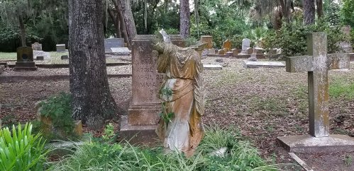 A weathered statue of a woman stands in a cemetery, surrounded by gravestones and overgrown greenery.