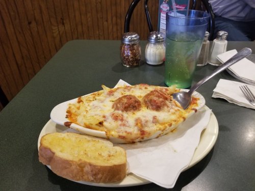 A plate of cheesy baked pasta with meatballs, served with a slice of garlic bread and a glass of green drink.