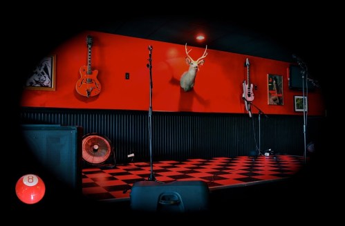 A stage with a checkered floor, red walls, guitars on display, and a mounted deer head. A pool ball is in the foreground.