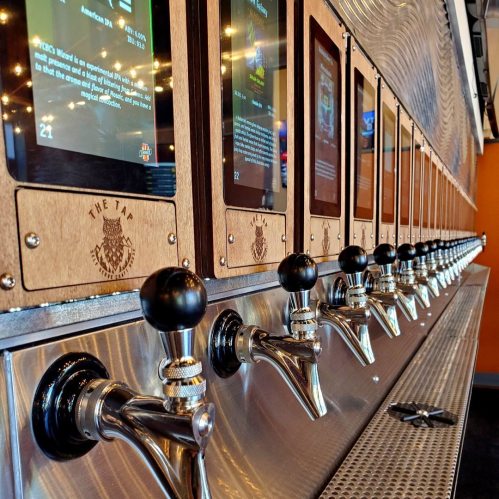 A row of beer taps with digital screens displaying information about each drink. Modern bar setting.