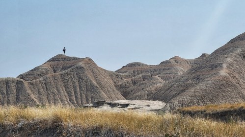 A person stands atop a rugged, brown hill, surrounded by layered rock formations and dry grass under a clear blue sky.