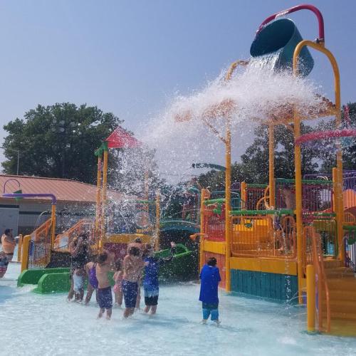 Children play in a water park, enjoying a colorful splash pad with water slides and a large water bucket overhead.