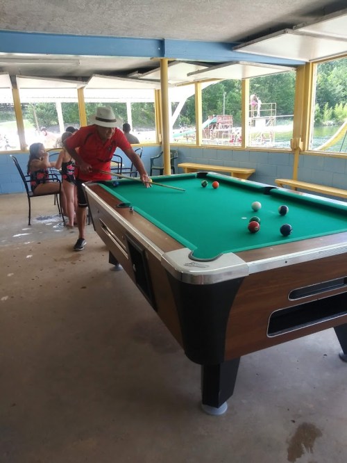 A man in a hat leans over a pool table, while a woman sits nearby in a casual setting.