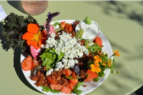 A colorful salad with greens, tomatoes, edible flowers, blue cheese, and a side of dressing on a white plate.