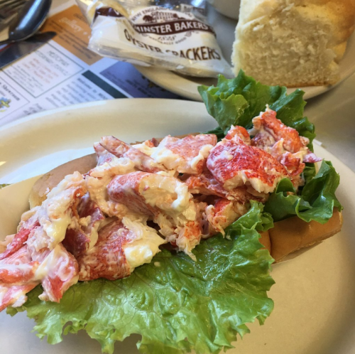 A lobster roll topped with lettuce on a plate, accompanied by a piece of bread and a packet of oyster crackers.