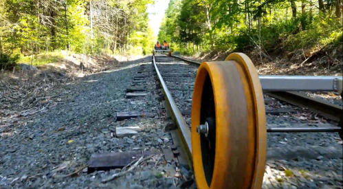 A close-up of a railway wheel on tracks, with trees lining the path and people in the distance.