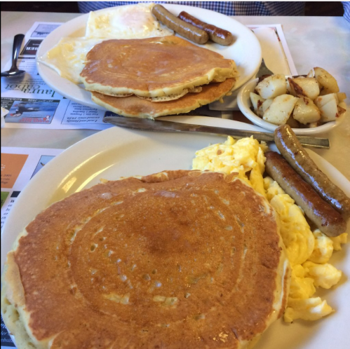 Two plates of breakfast featuring pancakes, scrambled eggs, sausage links, and a side of diced potatoes.