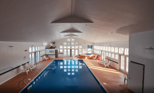 Indoor swimming pool with large windows, wooden deck, and lounge chairs, illuminated by natural light.