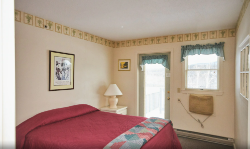 Cozy bedroom with a red bedspread, framed artwork, and a door leading to a balcony. Light-colored walls and curtains.