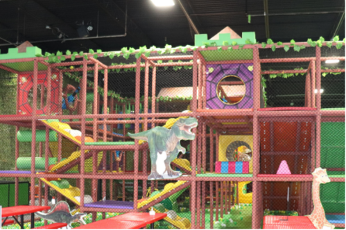 Colorful indoor playground with slides, climbing structures, and dinosaur decorations, surrounded by greenery.