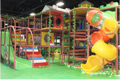 Colorful indoor playground with slides, climbing structures, and a large spiral slide, surrounded by greenery.