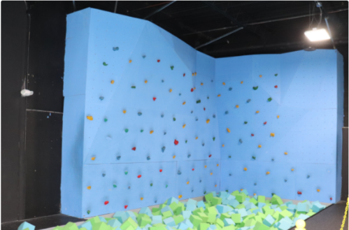 Indoor climbing wall with colorful holds and a foam pit below, set against a blue backdrop.