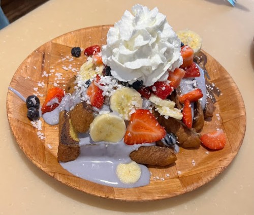 A wooden plate filled with a colorful dessert topped with whipped cream, strawberries, bananas, blueberries, and coconut.