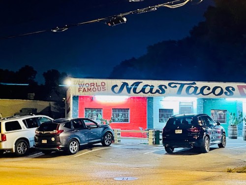 A brightly lit taco restaurant at night, featuring colorful walls and parked cars in front.