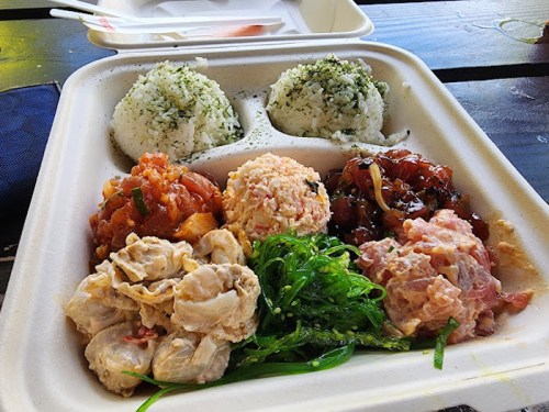 A takeout container with rice balls and various seafood dishes, including poke, seaweed salad, and marinated proteins.