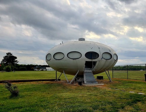 A futuristic, oval-shaped building with large windows, set on a grassy field under a cloudy sky.