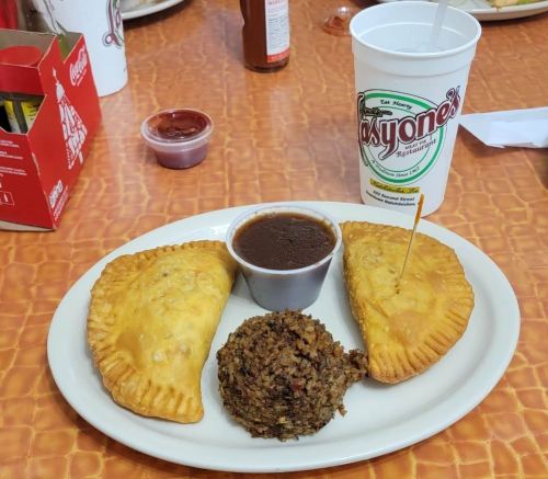 A plate with two golden empanadas, a serving of rice, and a cup of sauce, alongside a drink from a restaurant.