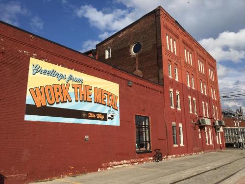 A red brick building with a large mural reading "Greetings from WORK THE METAL" and a clear blue sky above.
