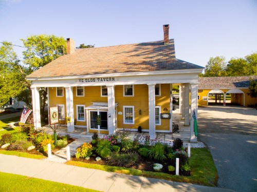 A yellow tavern with white columns, surrounded by greenery and flowers, featuring an American flag and a sign out front.