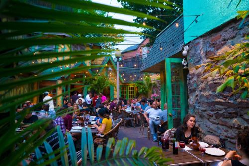 A lively outdoor restaurant scene with diners enjoying meals among greenery and string lights.