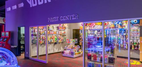 Brightly lit prize center filled with colorful toys and games behind glass displays in a fun entertainment venue.