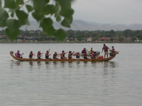 A group of people paddling a traditional dragon boat on a calm lake, surrounded by trees and distant hills.