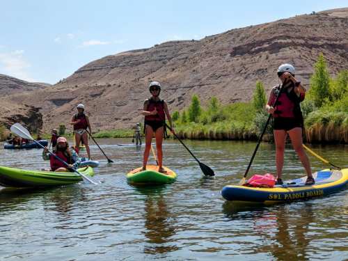 Group of people paddleboarding on a calm river, surrounded by rocky hills and greenery under a clear blue sky.