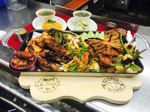 A large platter of grilled meats, vegetables, and sides, served with lime and salsa, on a wooden board.