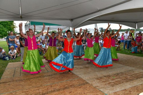 A group of dancers in colorful outfits perform on stage under a tent, with an audience watching in the background.