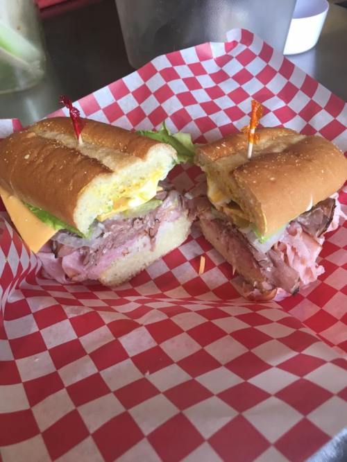 Two sandwiches on a red and white checkered paper, filled with meats, cheese, lettuce, and topped with toothpicks.