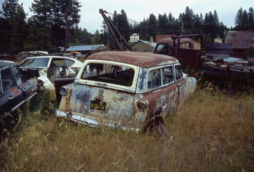 A rusty, vintage station wagon sits abandoned in a field, surrounded by other old cars and overgrown grass.