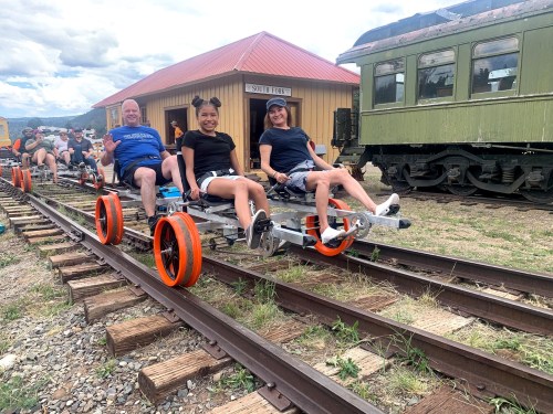 Two people sit on a rail bike on train tracks, with a vintage train and a building in the background.