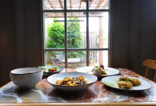 A table with four plates of food and a bowl, set against a window view of greenery and outdoor seating.