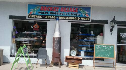 Exterior of a vintage resale shop with a rocket-themed sign, a large rocket display, and a chalkboard advertising sales.