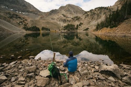 A person fishing by a serene mountain lake, surrounded by rocky terrain and reflecting peaks in the water.