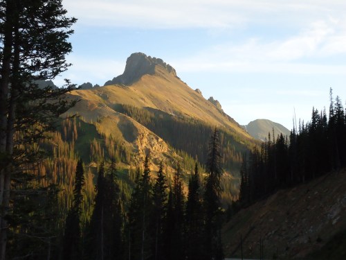 A rugged mountain peak rises above a forested landscape, illuminated by soft sunlight in the early evening.