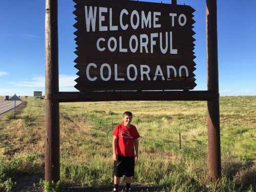 A boy stands in front of a large "Welcome to Colorful Colorado" sign in a grassy landscape.