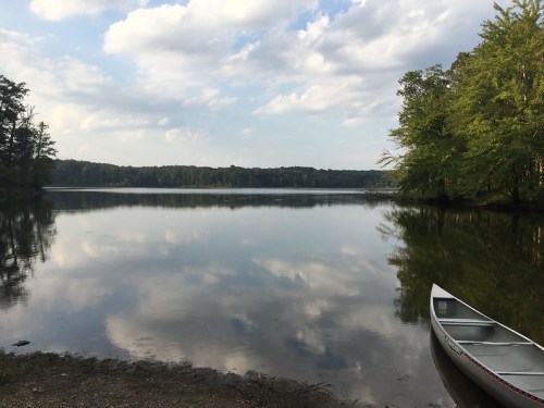 A calm lake reflects clouds and trees, with a canoe resting on the shore.