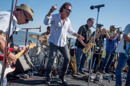 A lively band performs on stage by the water, featuring guitars, brass instruments, and energetic vocals.